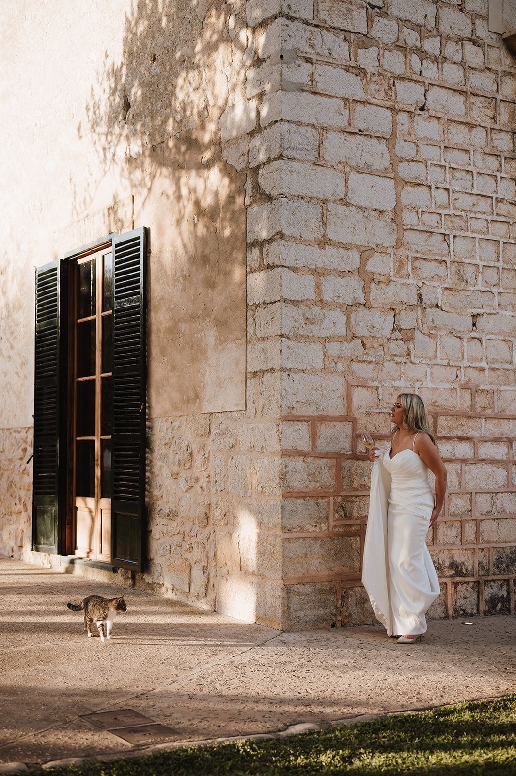 Bride in white gown standing by rustic stone building with a cat.