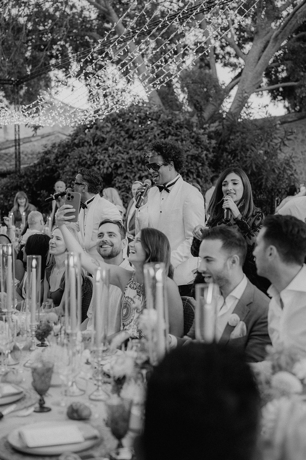 Wedding reception guests laughing and taking selfies under twinkling fairy lights at elegant dinner.