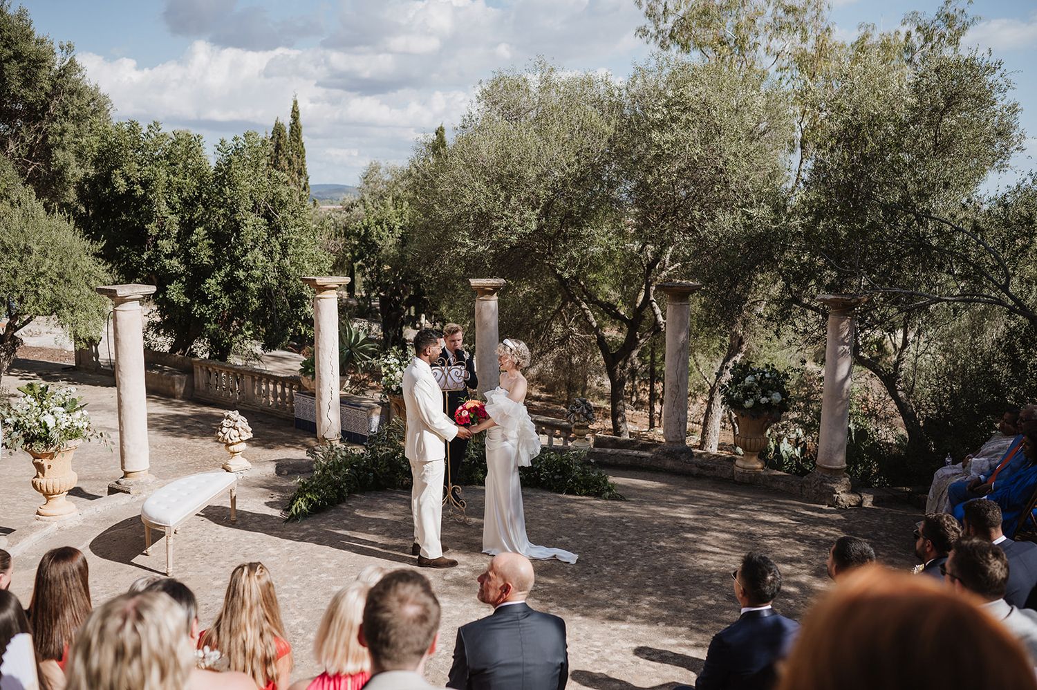 Outdoor wedding ceremony under olive trees with couple exchanging vows before seated guests.