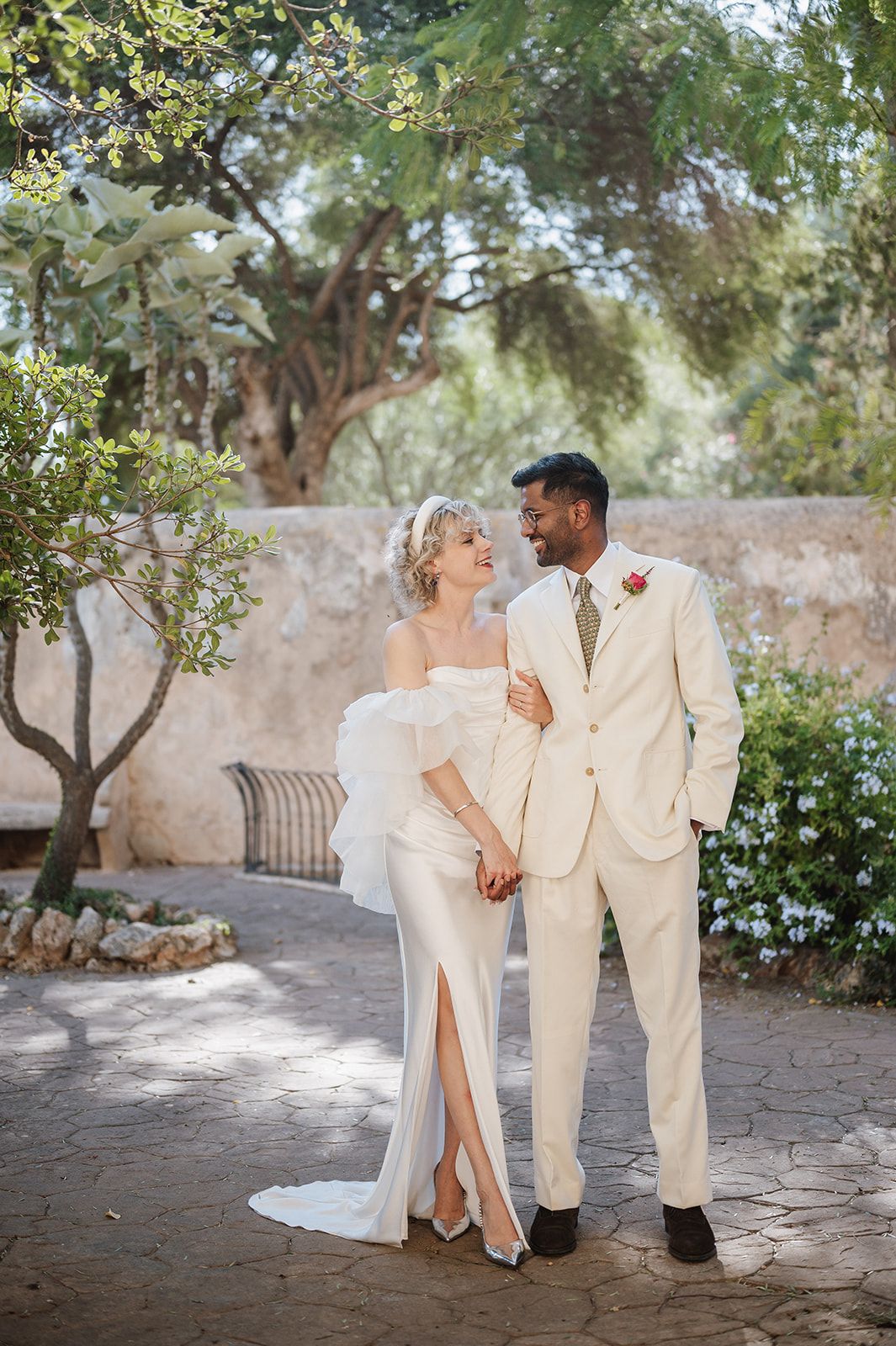 Interracial couple in wedding attire holding hands and smiling in a sun-dappled garden setting.