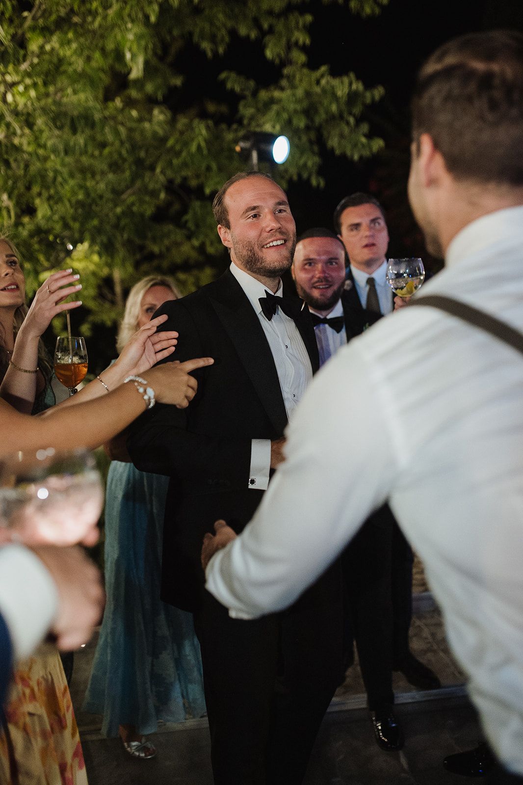 Smiling man in tuxedo greeting guests at elegant outdoor wedding reception under trees at night.