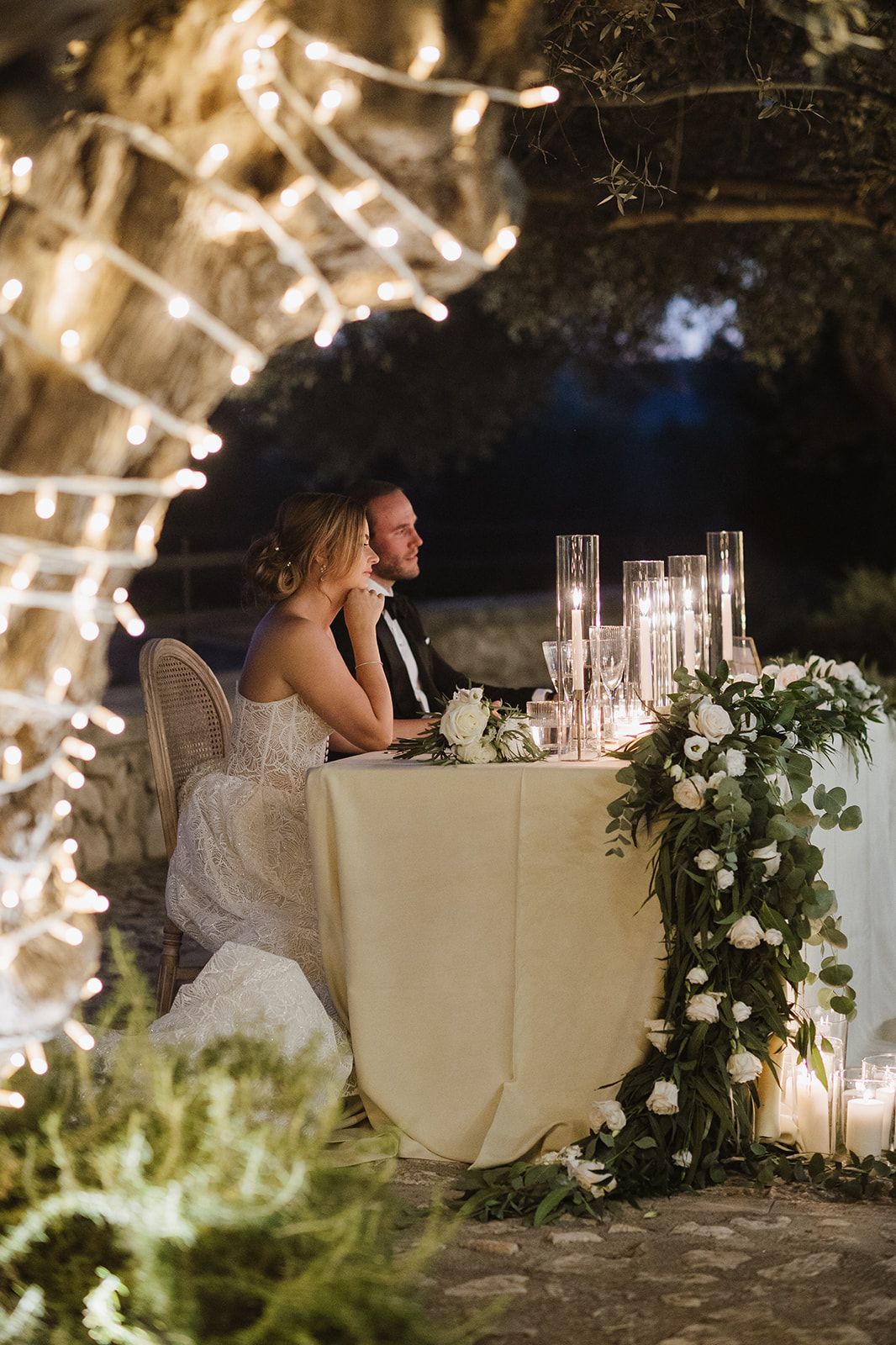 Bride and groom seated at romantic sweetheart table adorned with greenery garland and candles under fairy lights at night.
