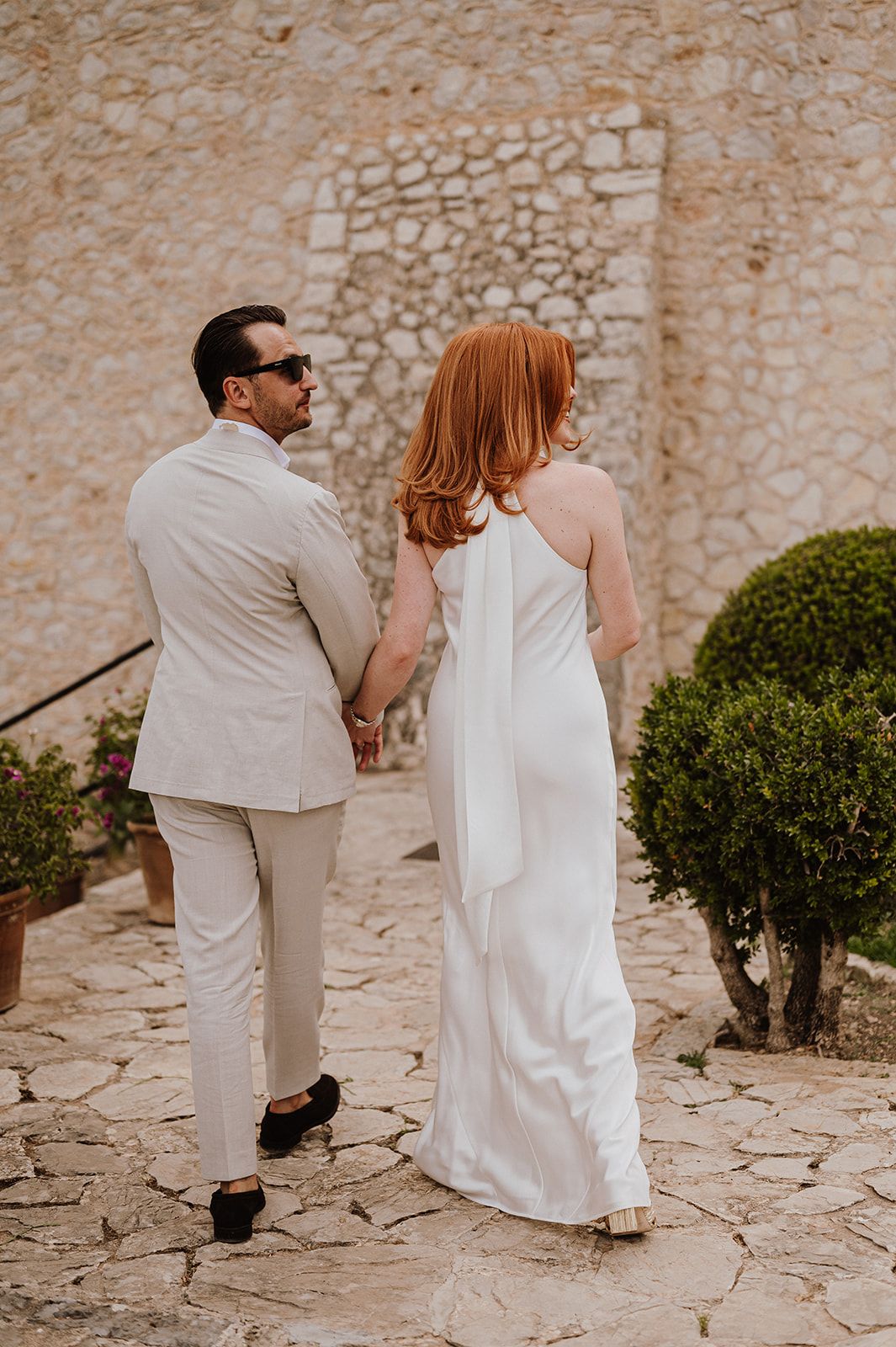 Newlywed couple walks hand in hand past rustic stone wall and trimmed hedges after ceremony.