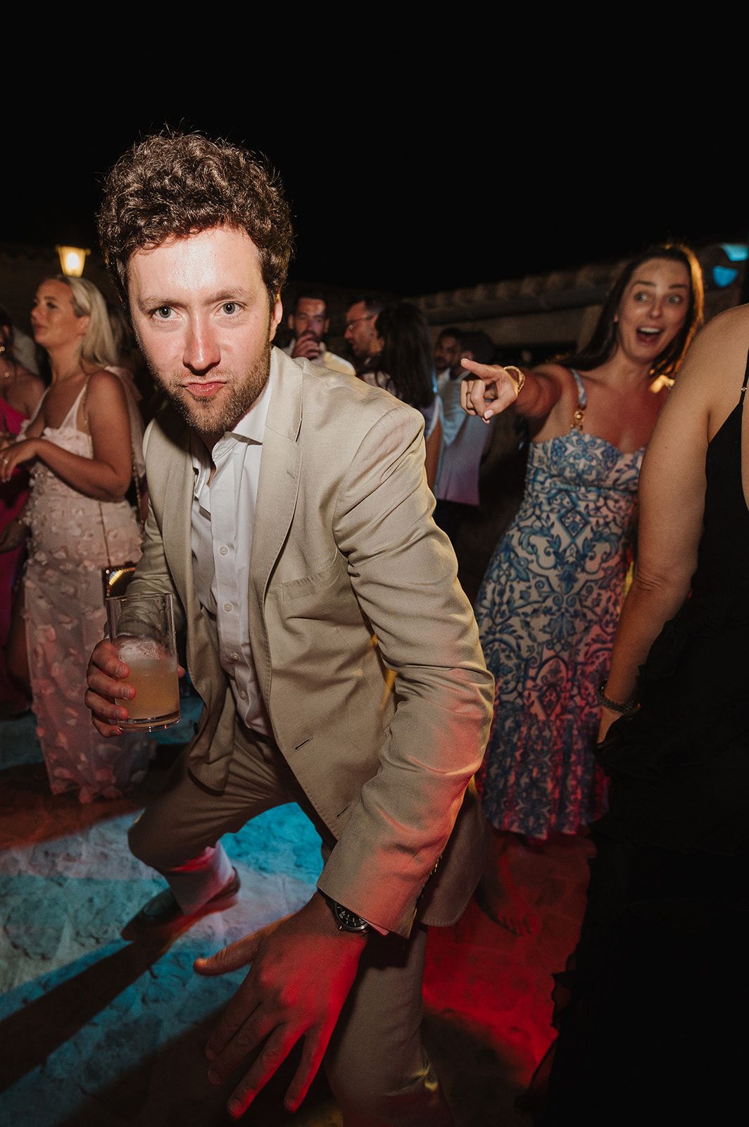 Curly-haired bearded man in tan suit dancing at a lively outdoor wedding reception.