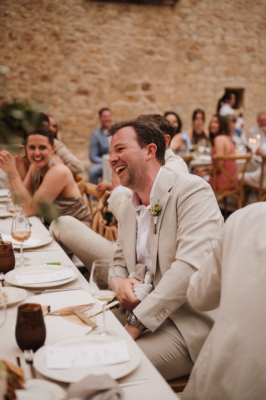 Groom in beige suit laughing during wedding reception speeches at a beautifully decorated dinner table.