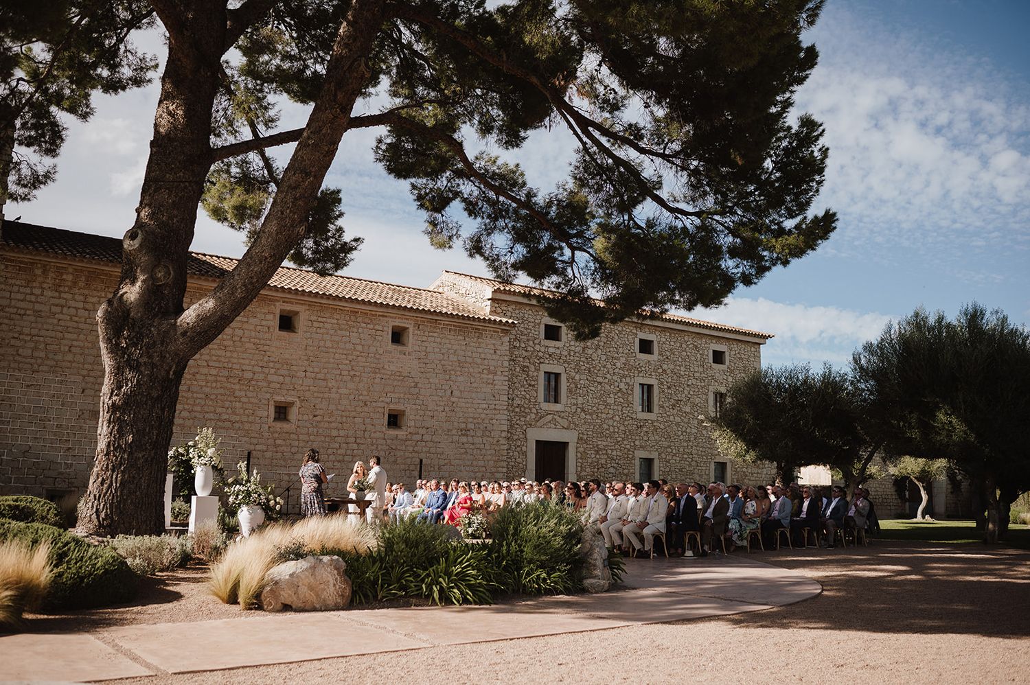 Outdoor wedding ceremony with guests gathered in front of a rustic stone building on a sunny day.