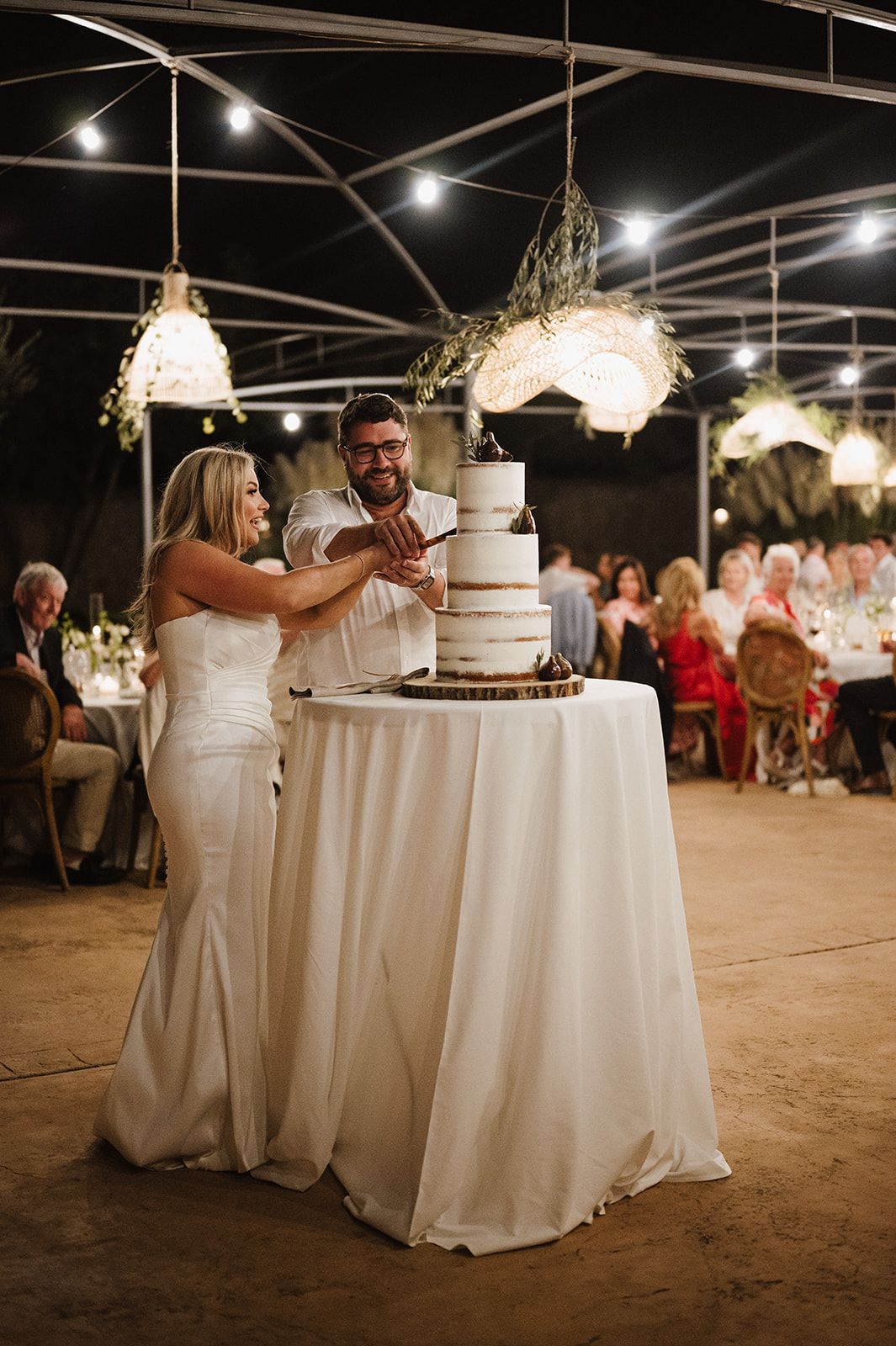 Bride and groom cutting white tiered wedding cake under string lights at evening reception.