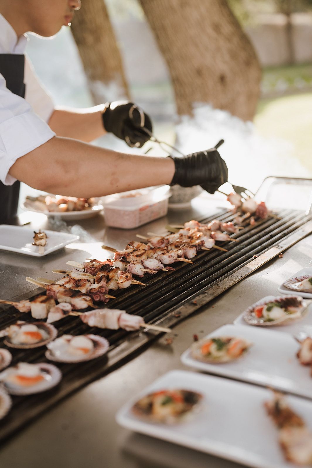Chef in black gloves arranging grilled seafood skewers and scallops on outdoor wedding grill.