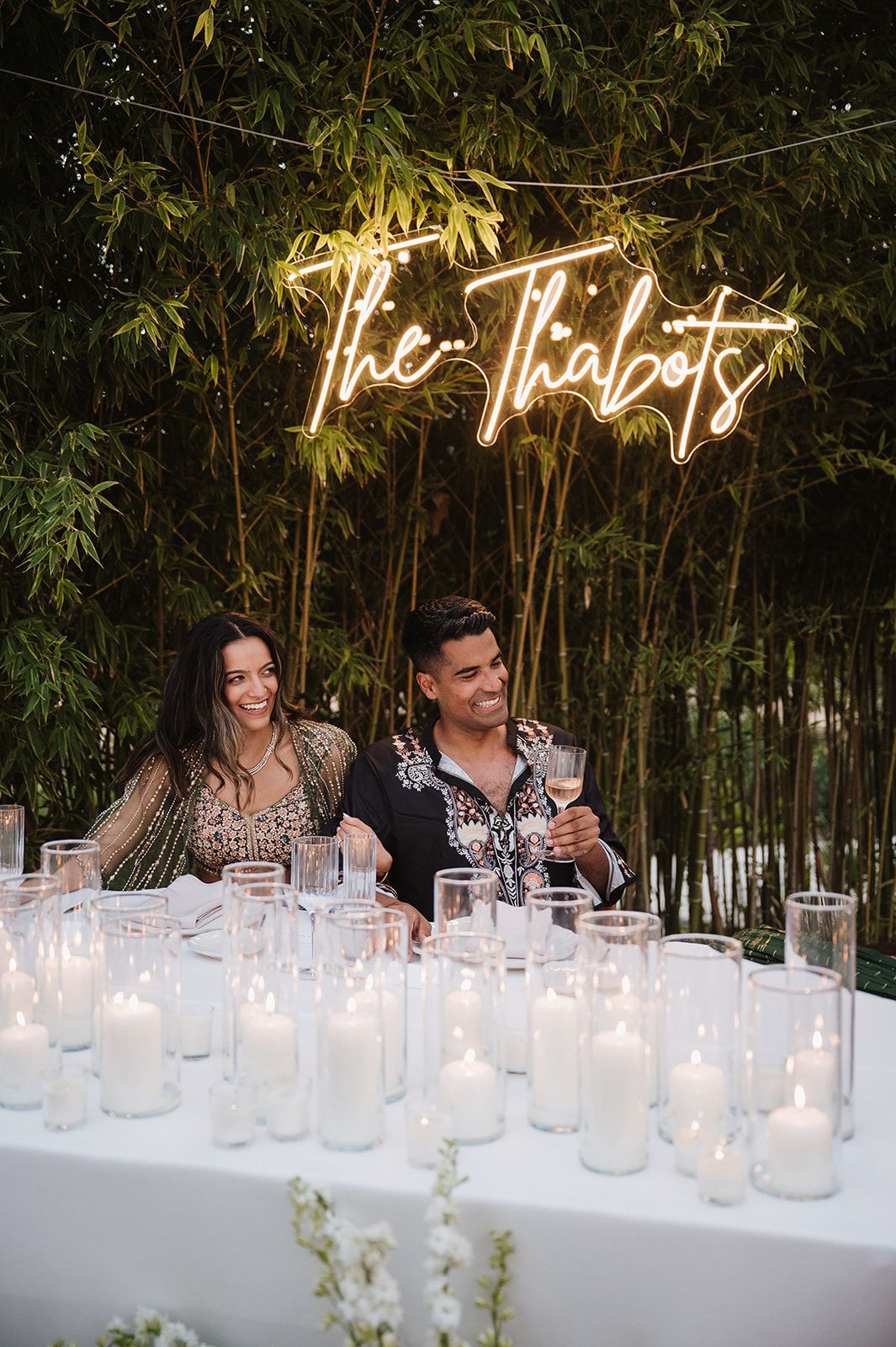 Couple smiles at candlelit wedding reception table under glowing neon sign reading The Thabots, surrounded by bamboo.