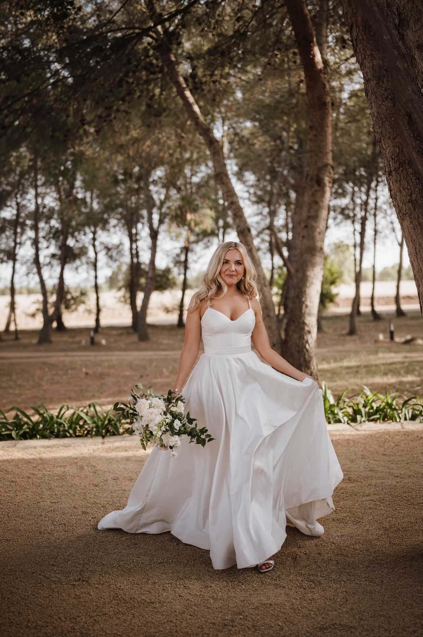 Elegant bride in a flowing white satin gown holds a white bouquet while posing among tall trees.