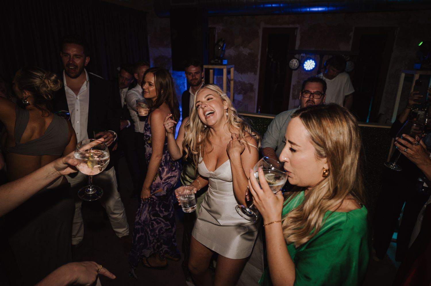 Blonde bride in white mini dress dances joyfully with wedding guests at vibrant indoor wedding after-party.
