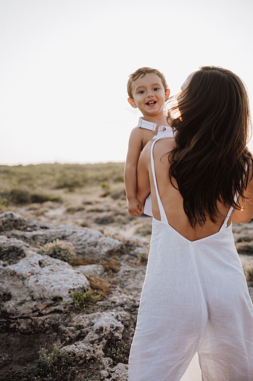 Mother in white backless dress holding smiling toddler on rocky sunlit landscape.