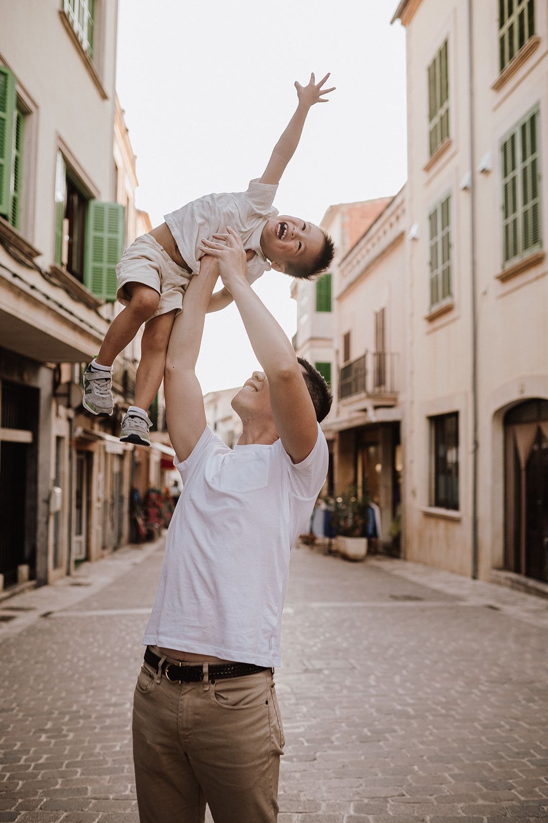 Father lifting happy child high in the air on a cobblestone European street.