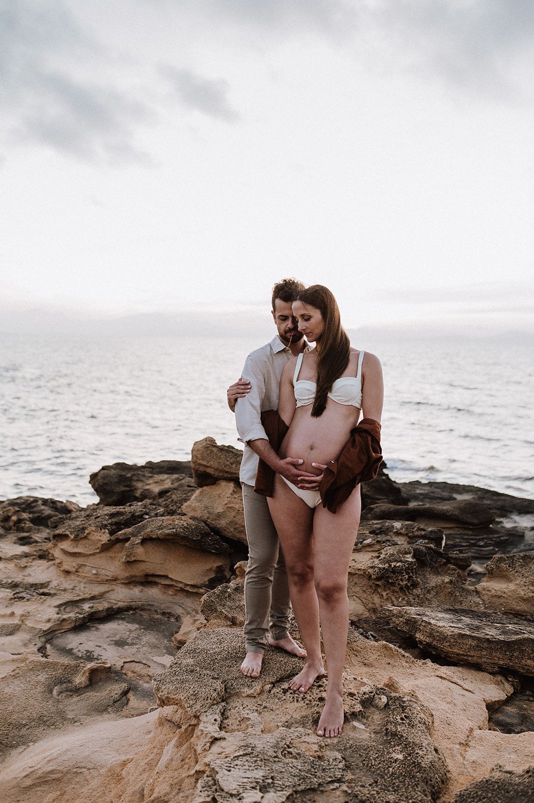 Pregnant couple embracing on rocky coastal cliffs, woman in white bikini with partner holding her baby bump by the ocean.