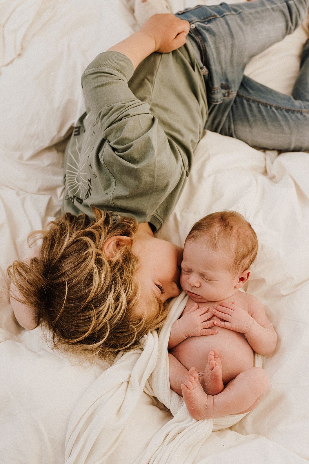 Young child lying on white bedding next to sleeping newborn baby, sharing a tender sibling moment.