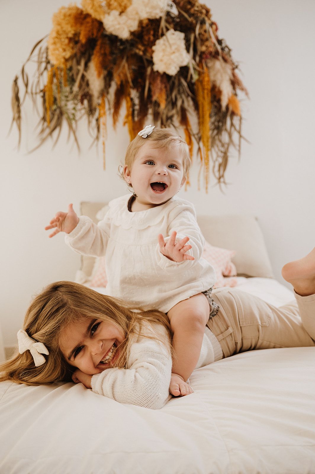 Laughing baby sitting on older sisters back on a white bed beneath a dried floral arrangement.
