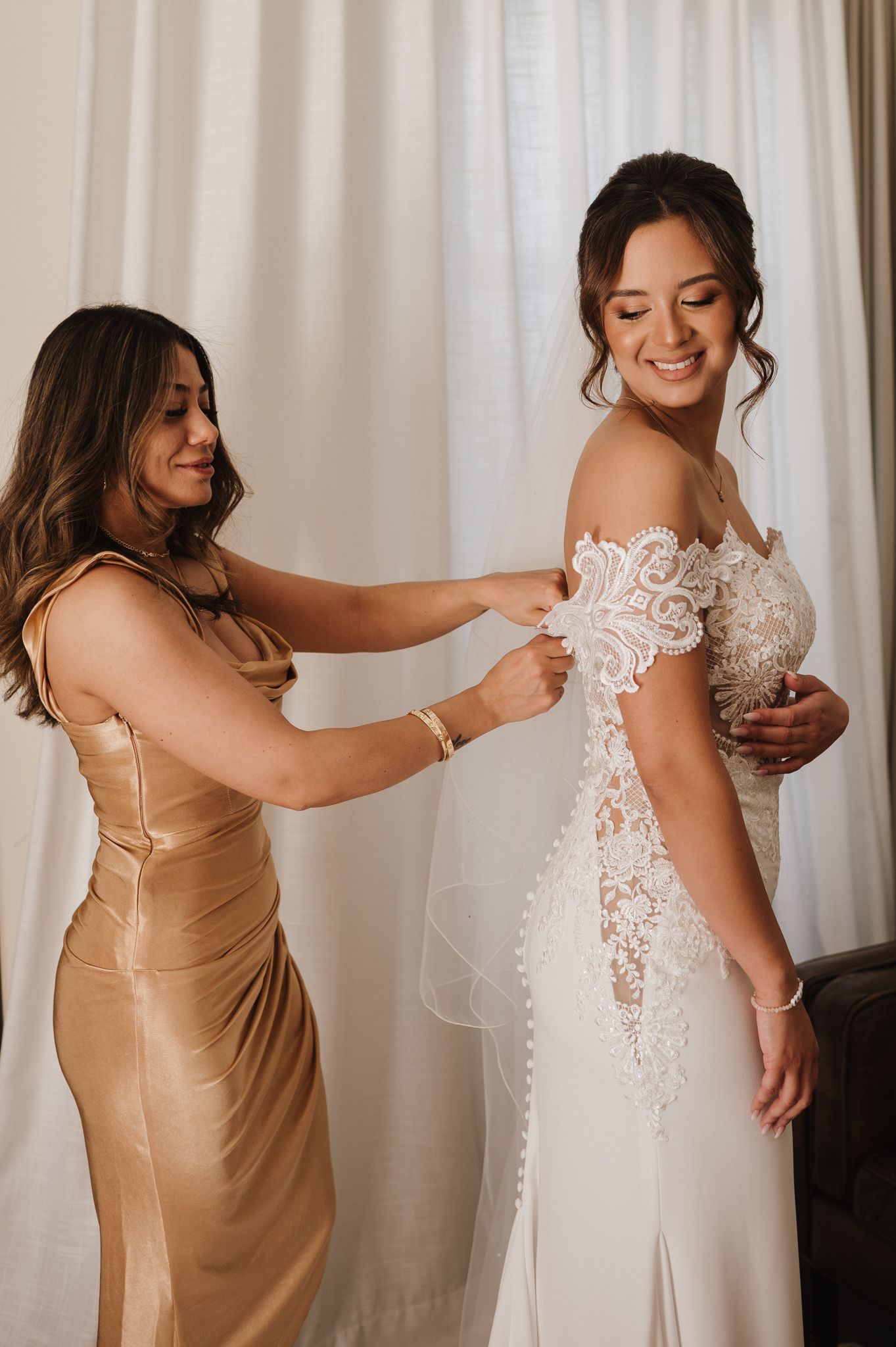 Bridesmaid in gold dress helping bride with lace wedding gown buttons before the ceremony.