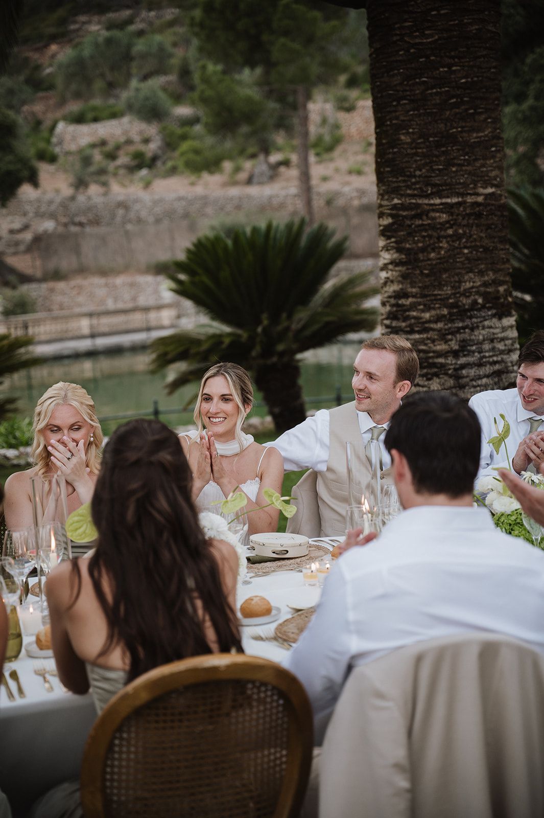 Bride and wedding guests laughing joyfully at outdoor reception table with mountain and tropical garden backdrop.