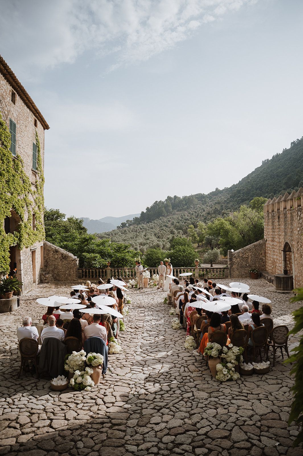 Wedding guests seated on cobblestone courtyard with parasols during an outdoor ceremony at a stone villa.