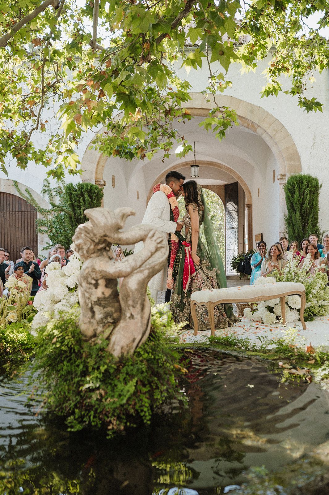 Bride and groom sharing first kiss at lush garden Hindu wedding ceremony by pond.