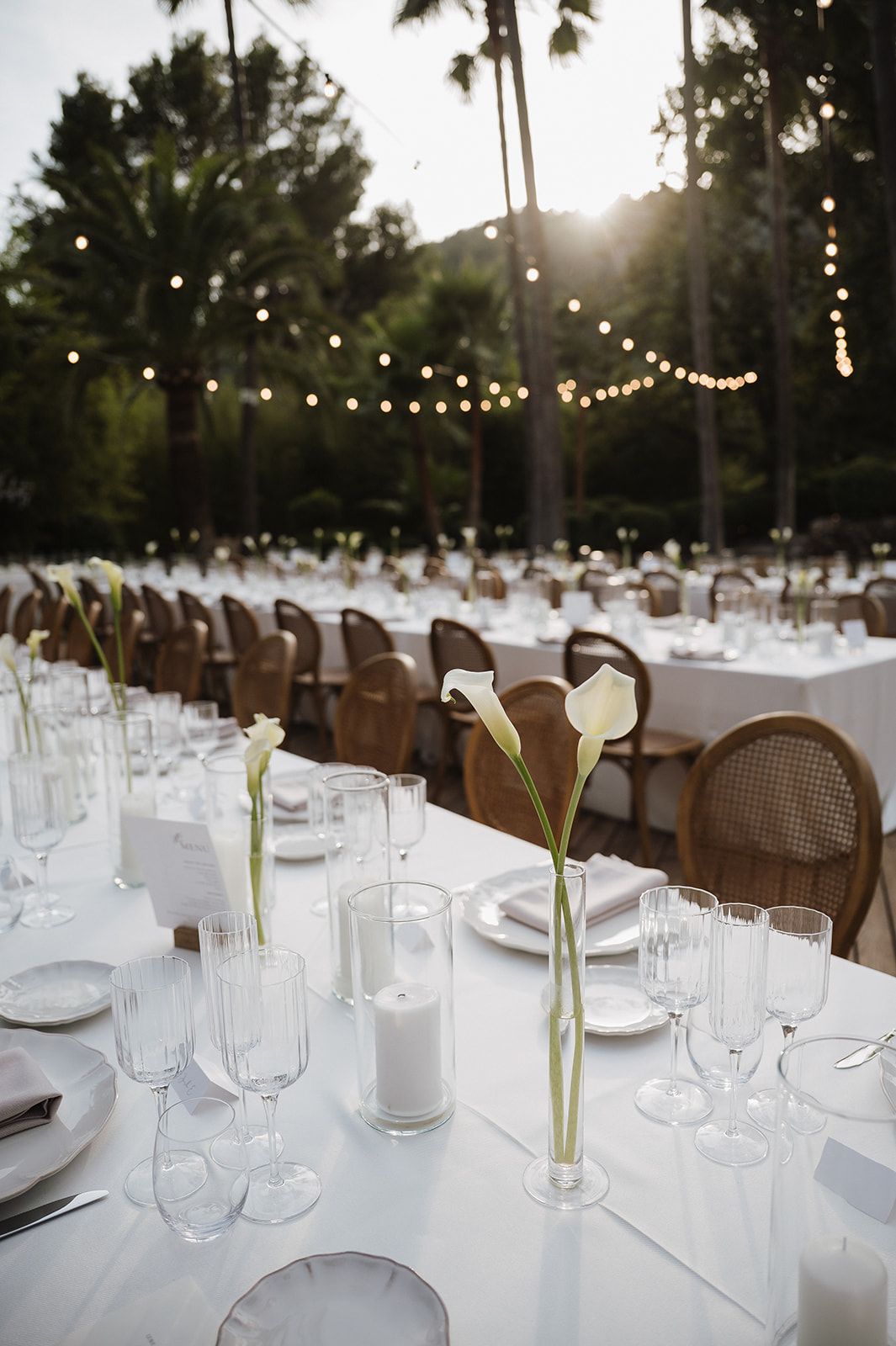 Elegant outdoor wedding reception with white tablecloths, calla lilies, and string lights at dusk.