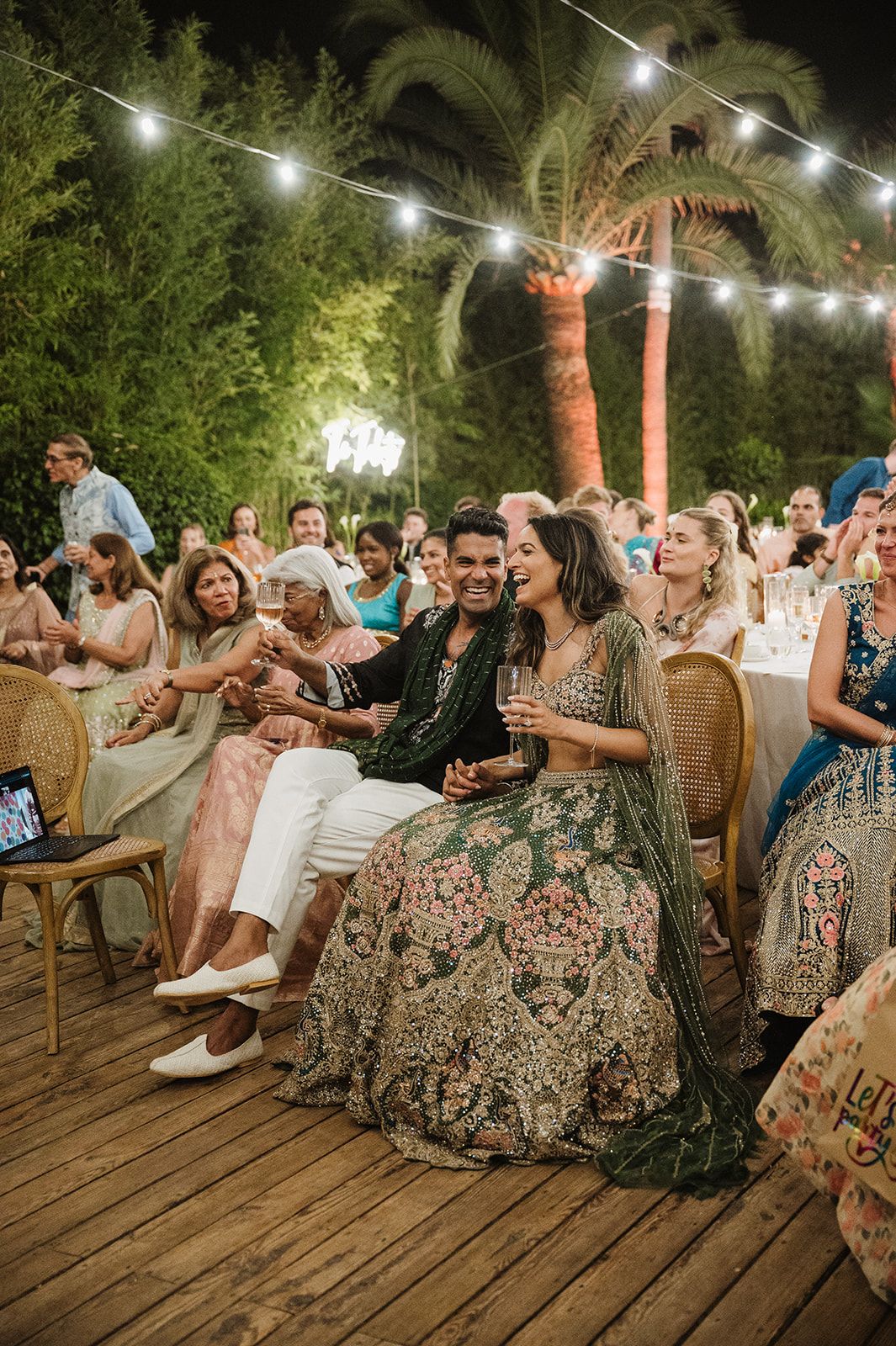 Indian bride and groom in embroidered green outfits laughing together at outdoor evening reception.