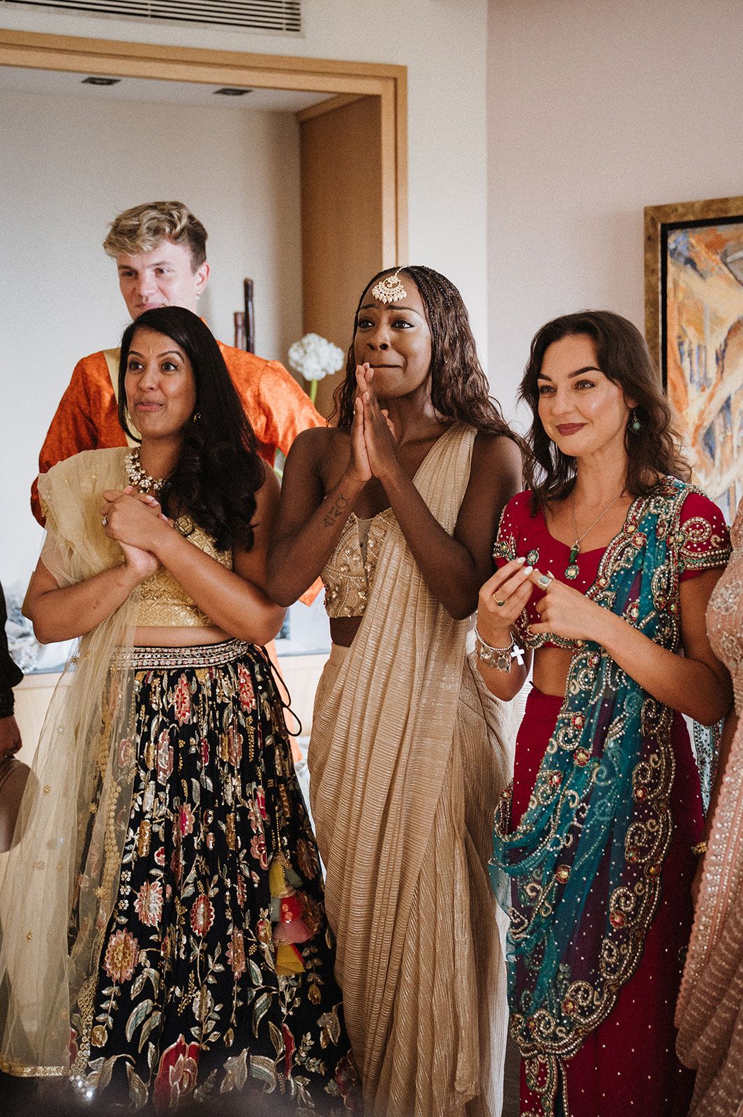 Women in traditional Indian attire smile and clap during a festive cultural celebration indoors.