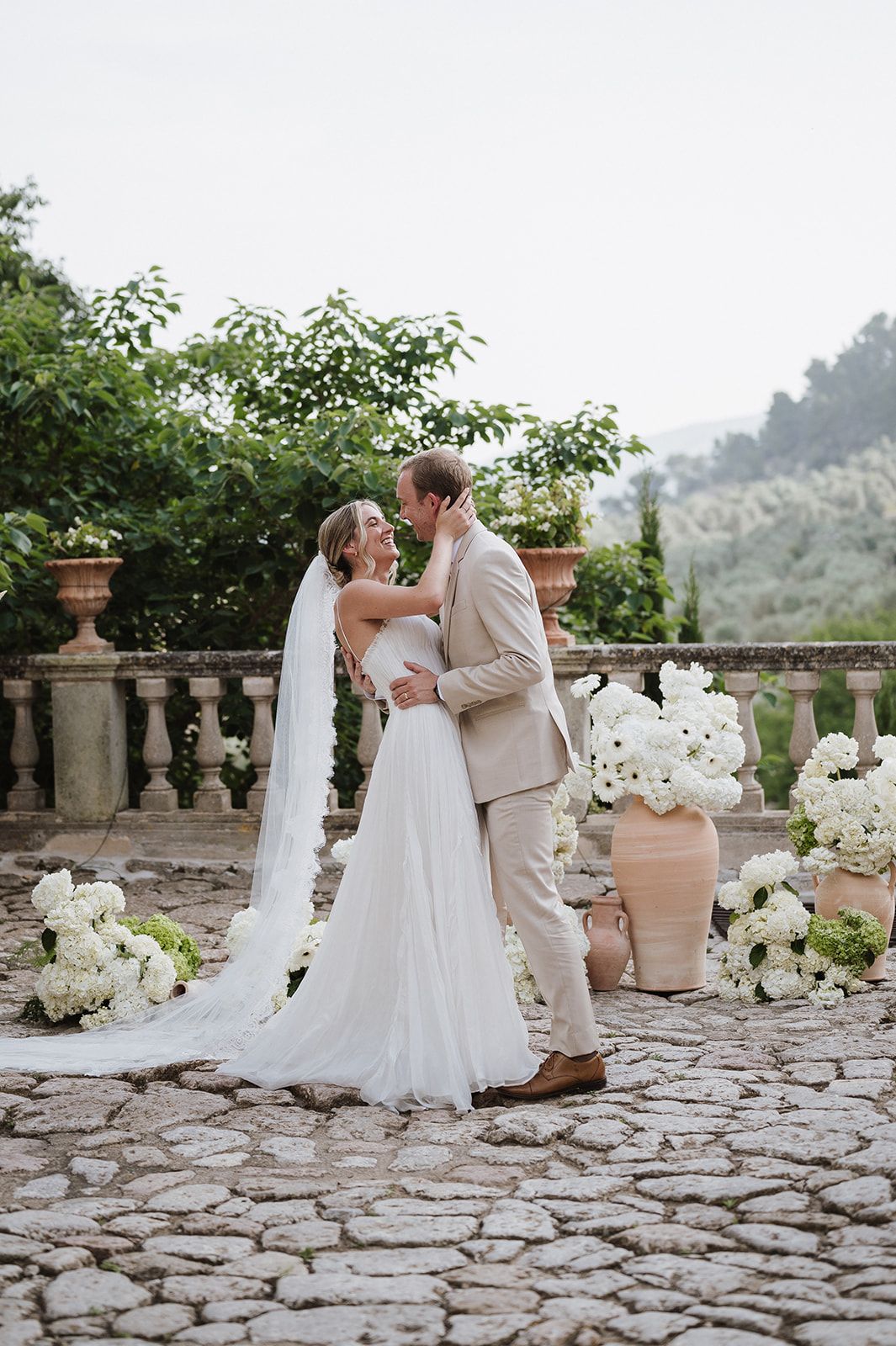 Bride and groom share romantic kiss on cobblestone terrace with white floral arrangements and veil.