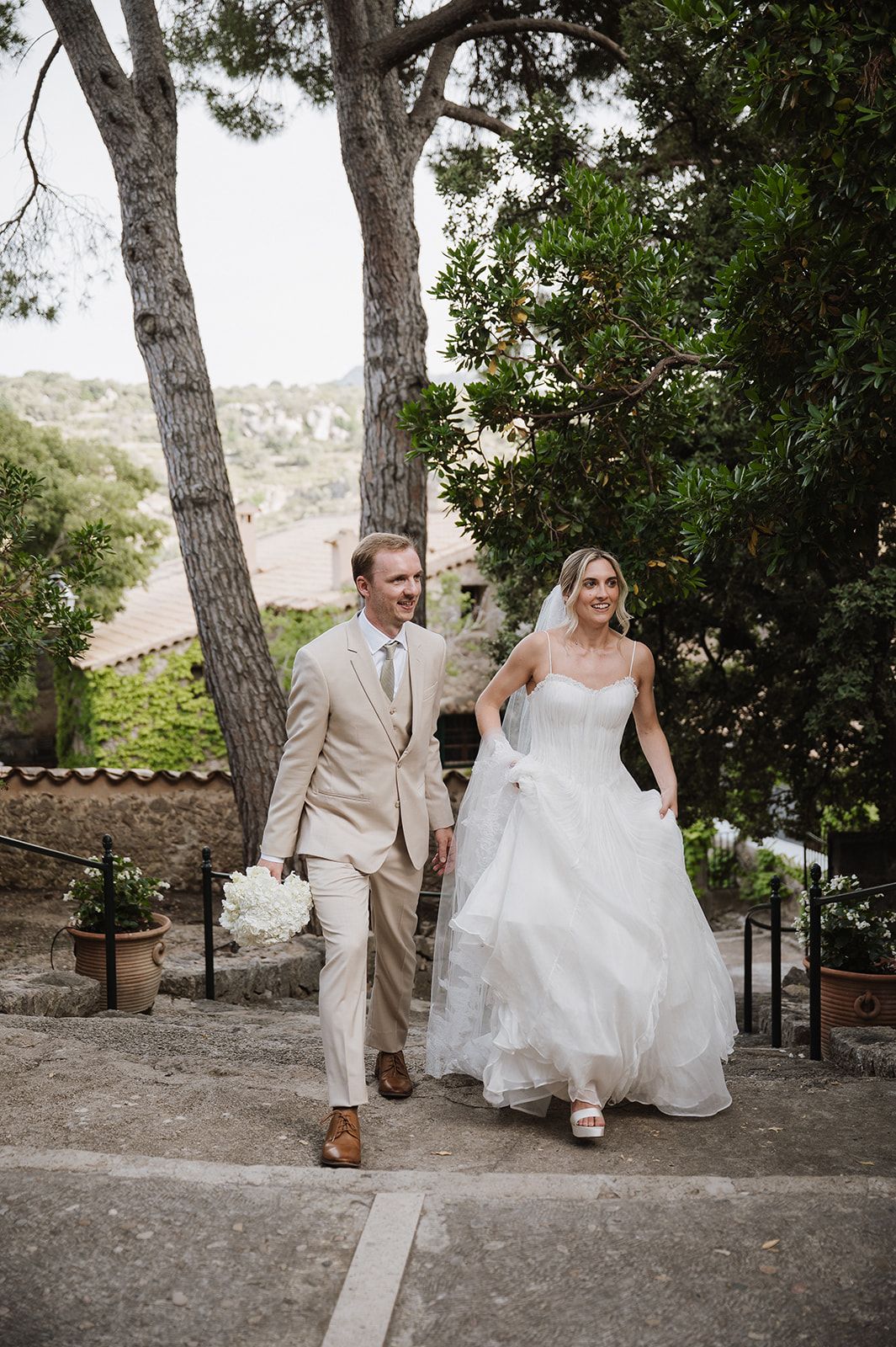 Smiling bride and groom in beige suit walk hand-in-hand down stone steps surrounded by Mediterranean greenery.