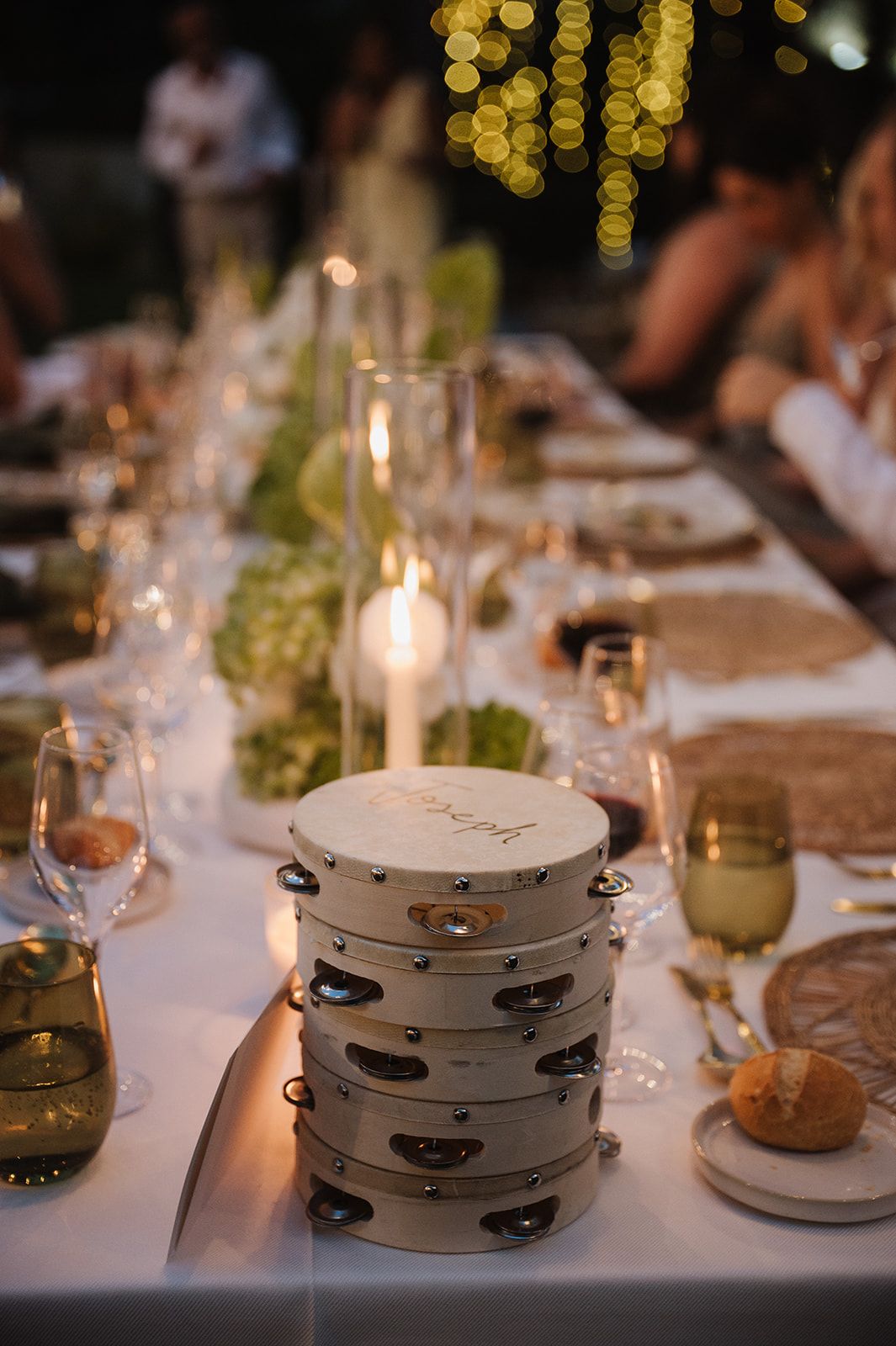 Elegant wedding reception table with stacked tambourines, candles, glassware, and greenery centerpieces under warm string lights.