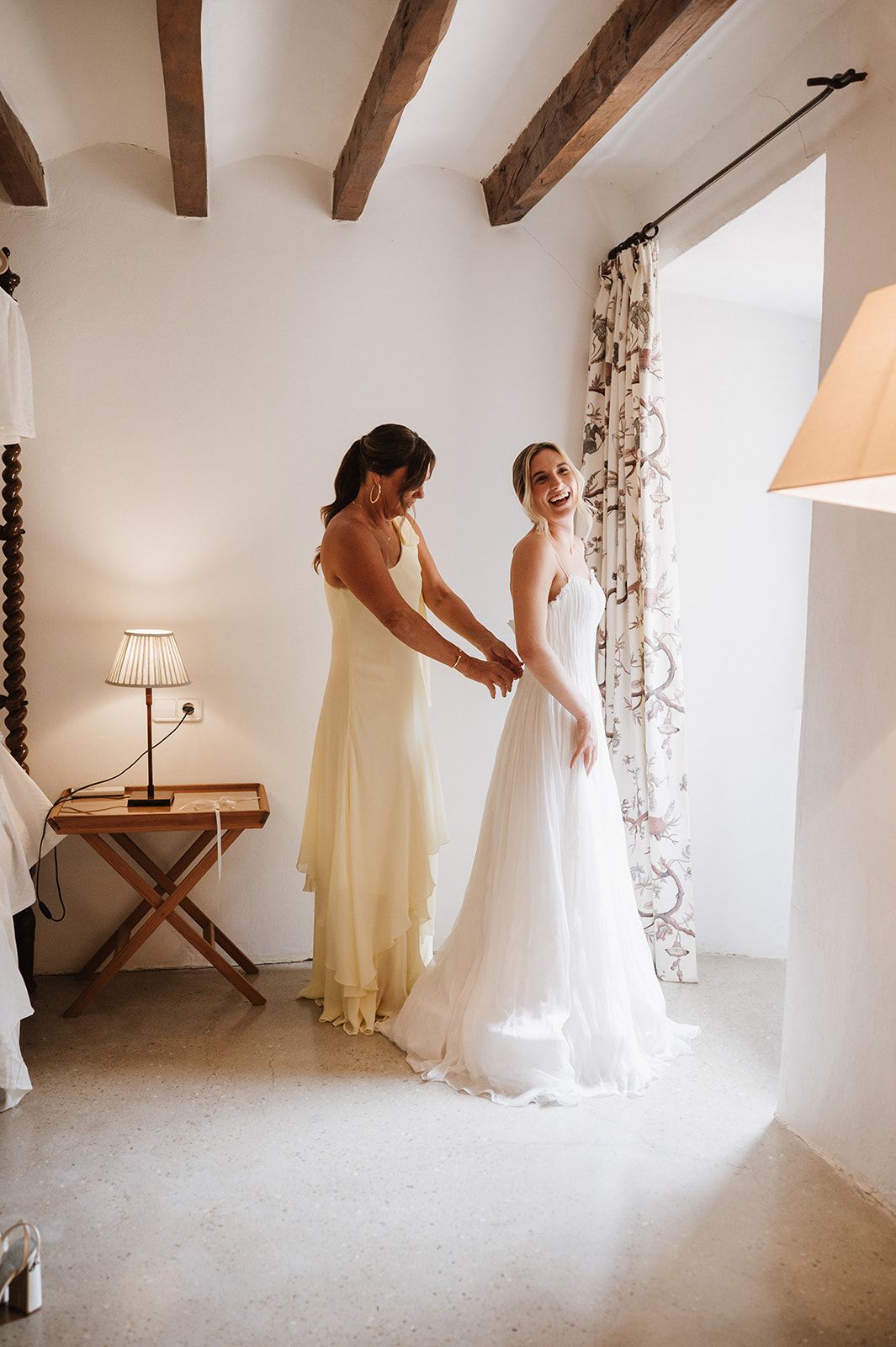 Bridesmaid in yellow dress helping smiling bride into white wedding gown in bright rustic room with wooden ceiling beams.