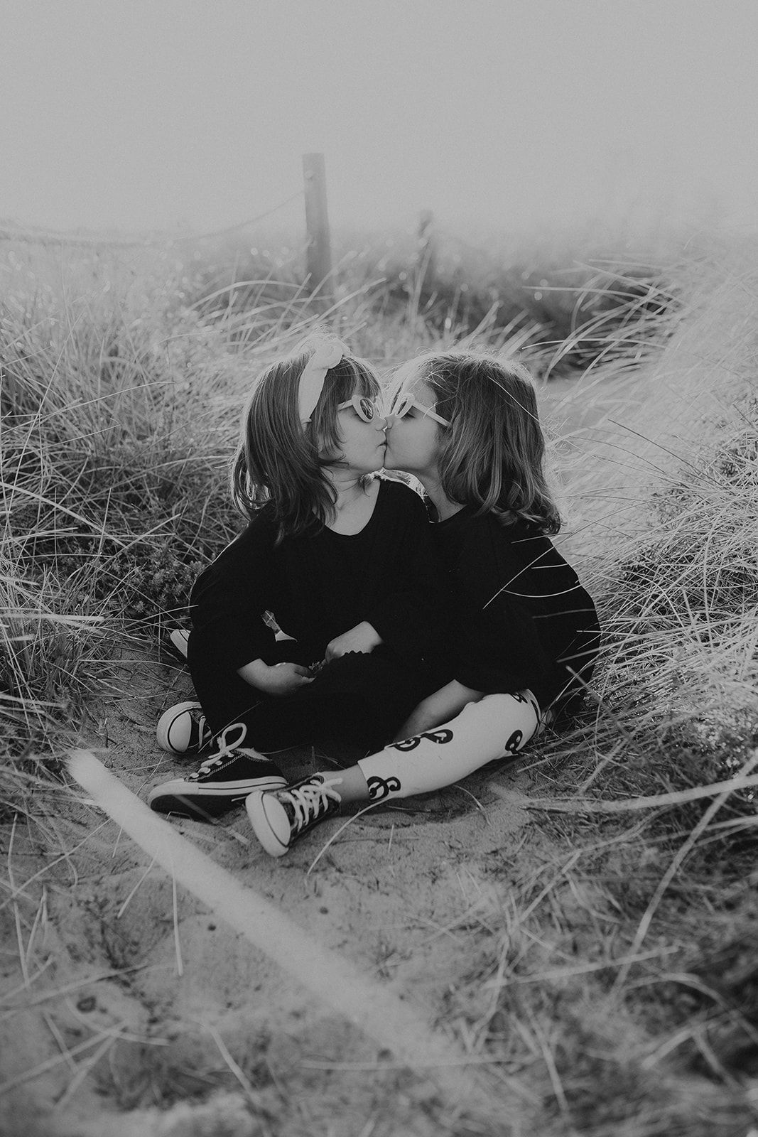 Two young children sharing a kiss while sitting in tall grass, captured in black and white photography.
