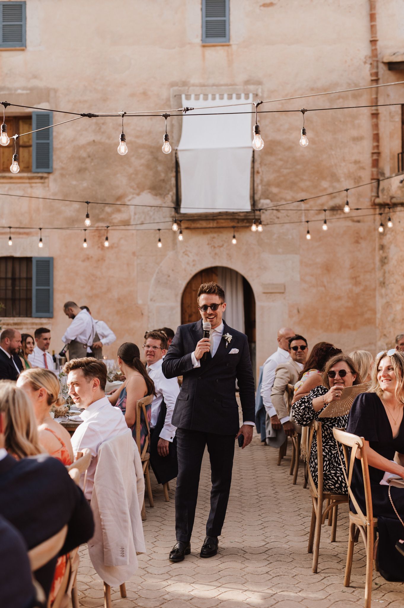 Groom in black suit giving speech at outdoor wedding reception in rustic Italian courtyard with string lights.
