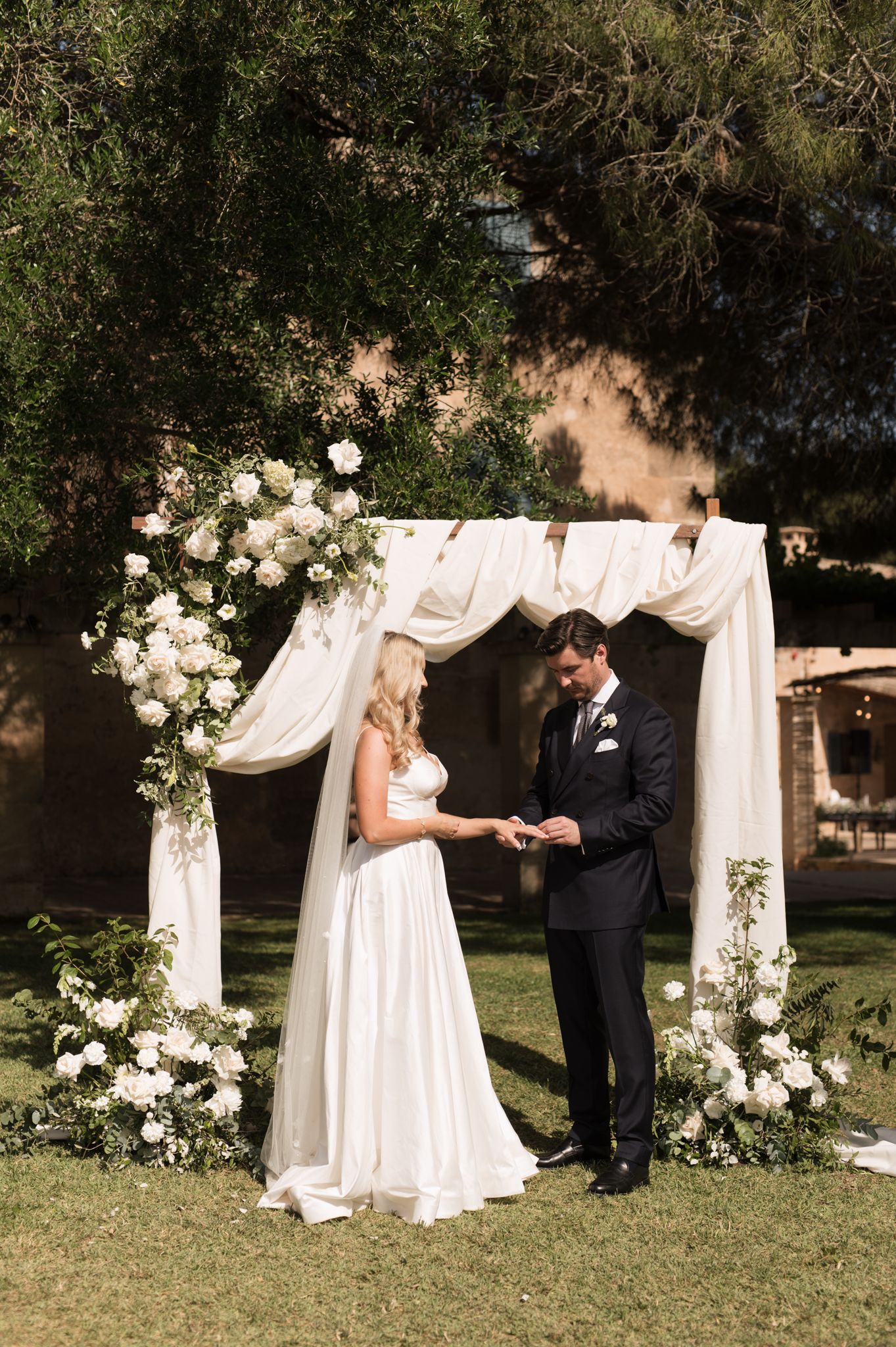 Groom places ring on bride's finger at floral arch altar with white roses and draped fabric outdoors.
