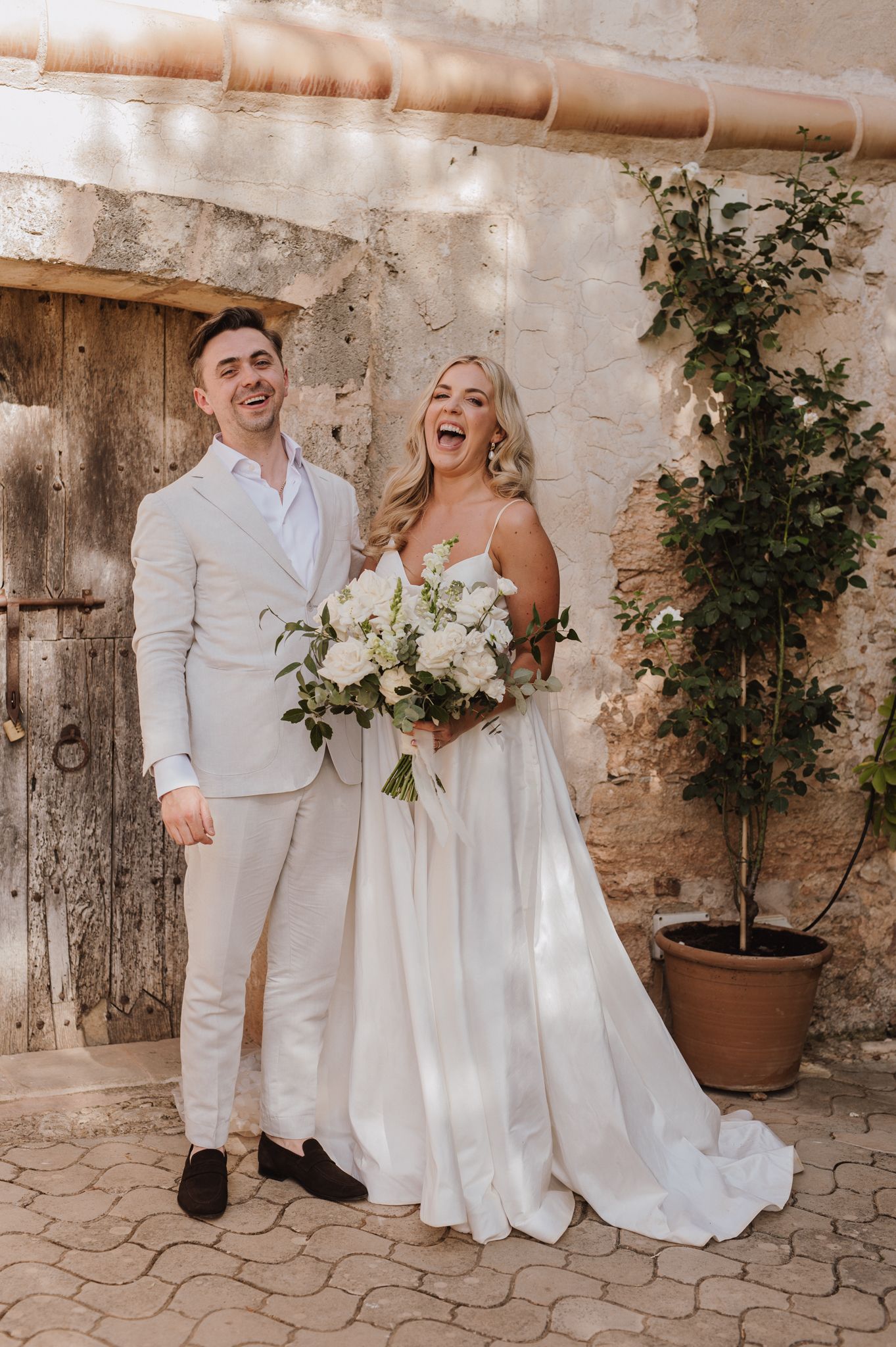 Laughing bride and groom posing together outside rustic stone building, bride holding white floral bouquet.