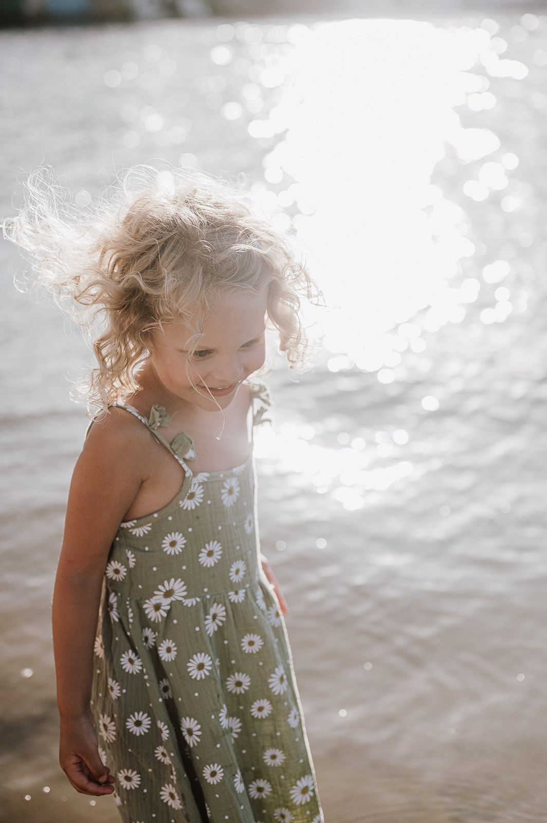 Curly blonde girl in daisy sundress smiling near sparkling sunlit water.