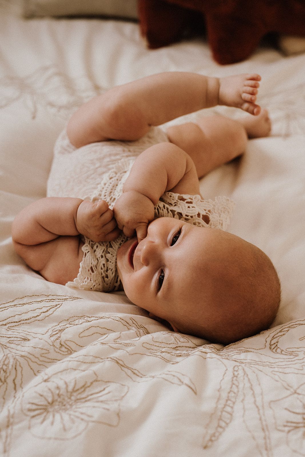 A happy baby in a white lace onesie lying on a floral patterned bed, smiling and holding their feet up.
