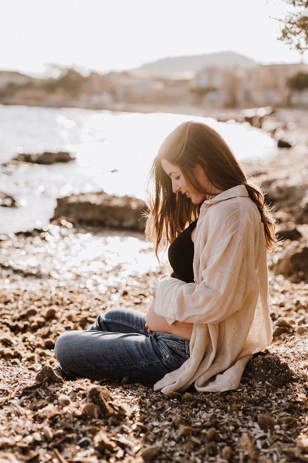 Pregnant woman sitting on rocky beach at golden hour, cradling baby bump in open linen shirt and jeans.