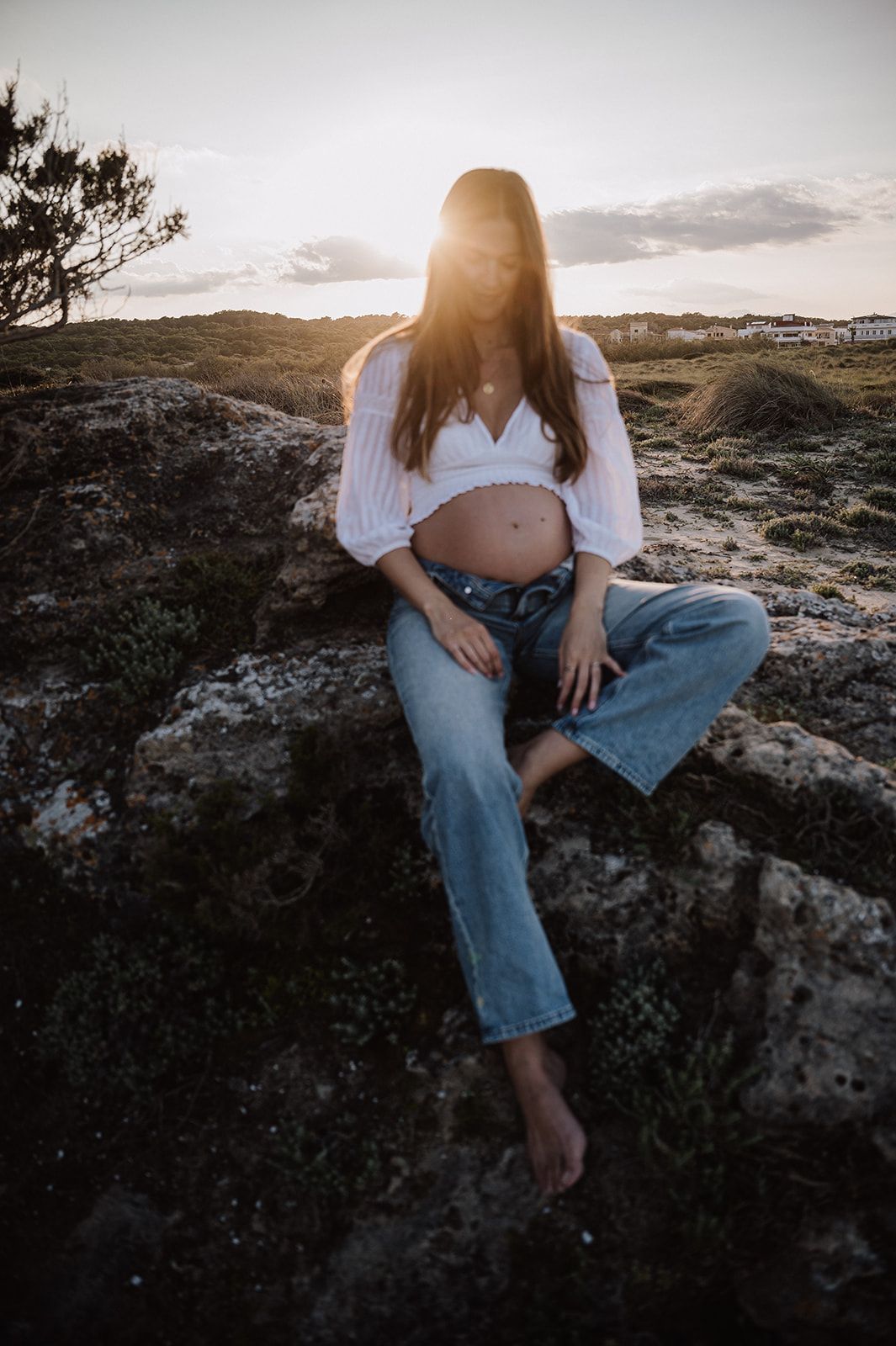 Pregnant woman sitting on rocks at sunset wearing white crop top and jeans, showing her baby bump outdoors.