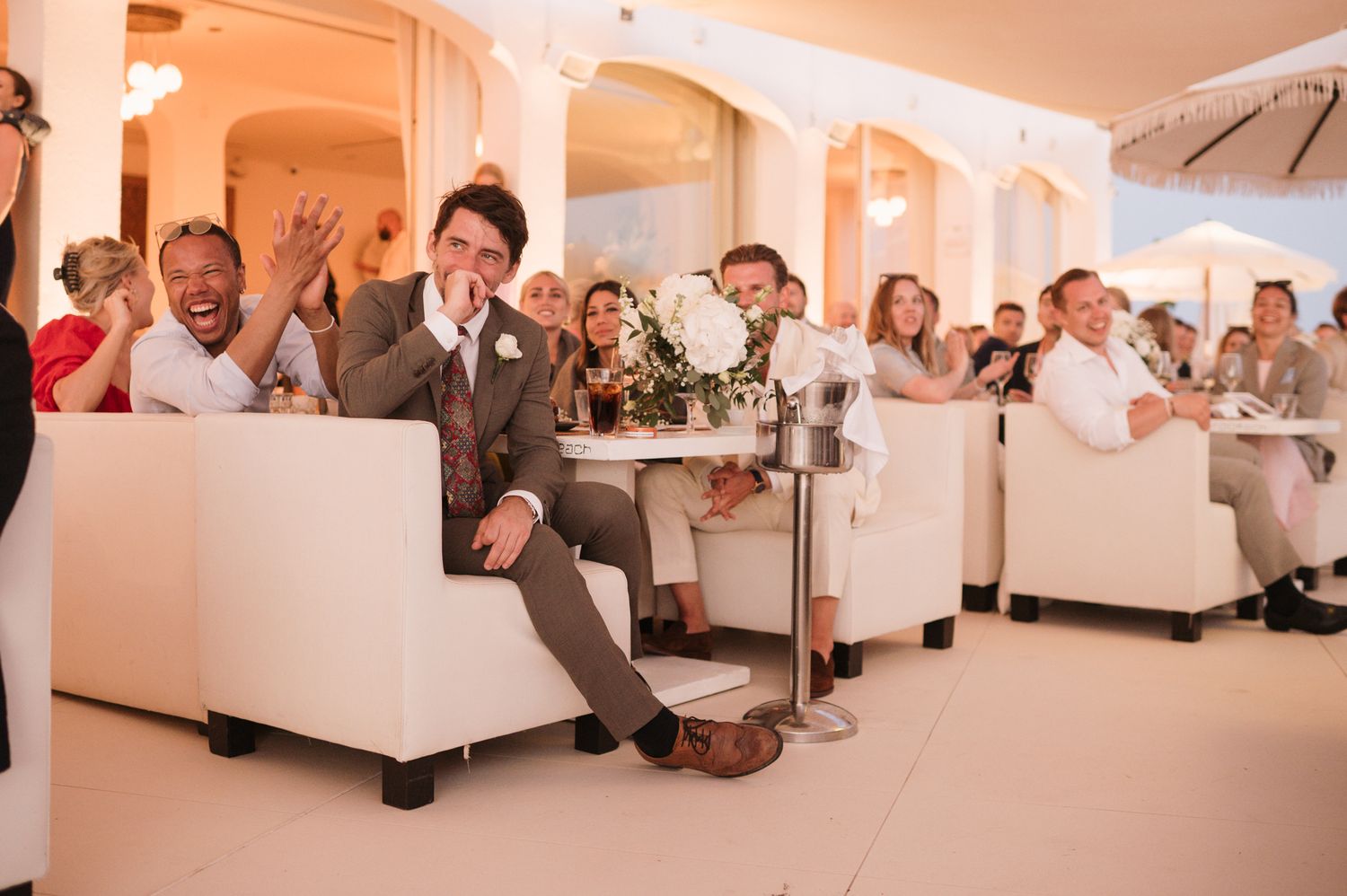 Guests laugh and react joyfully during wedding speeches in elegant white venue with floral arrangements.