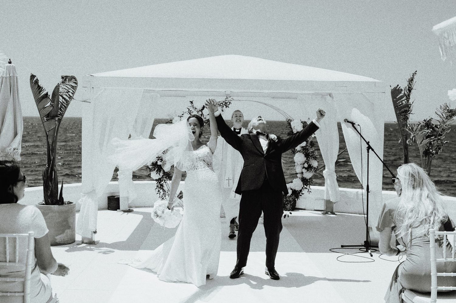 Joyful newlyweds raise arms in celebration at outdoor seaside wedding ceremony.