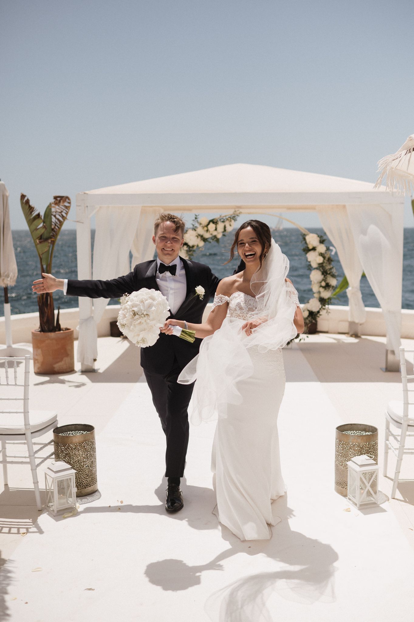 Laughing groom lifts bride in white gown at seaside wedding venue with white floral arch.
