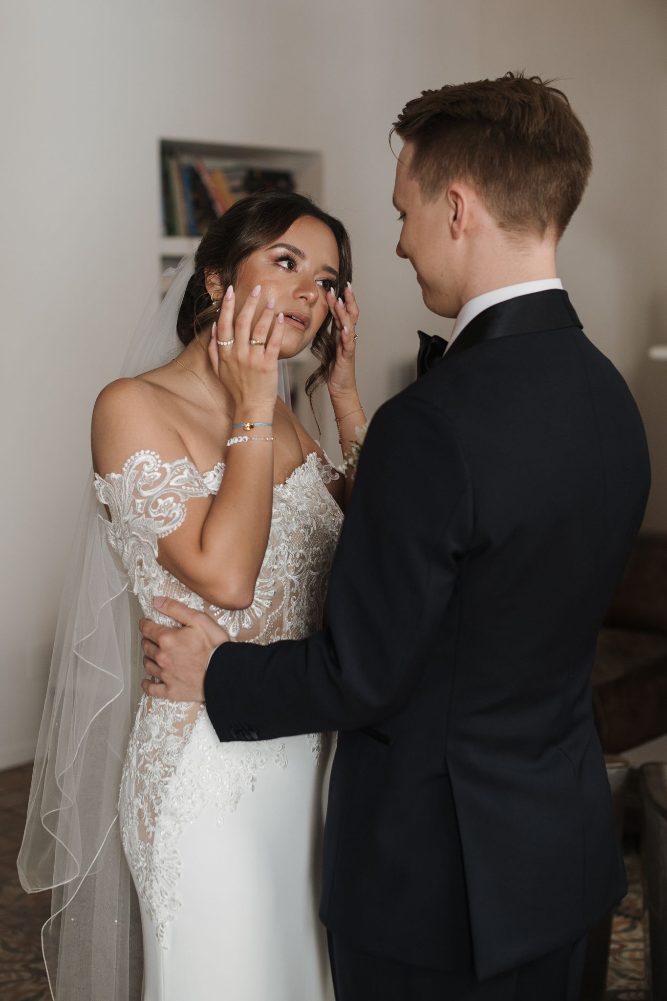 Emotional bride touches her face as groom tenderly holds her waist during intimate first look moment.