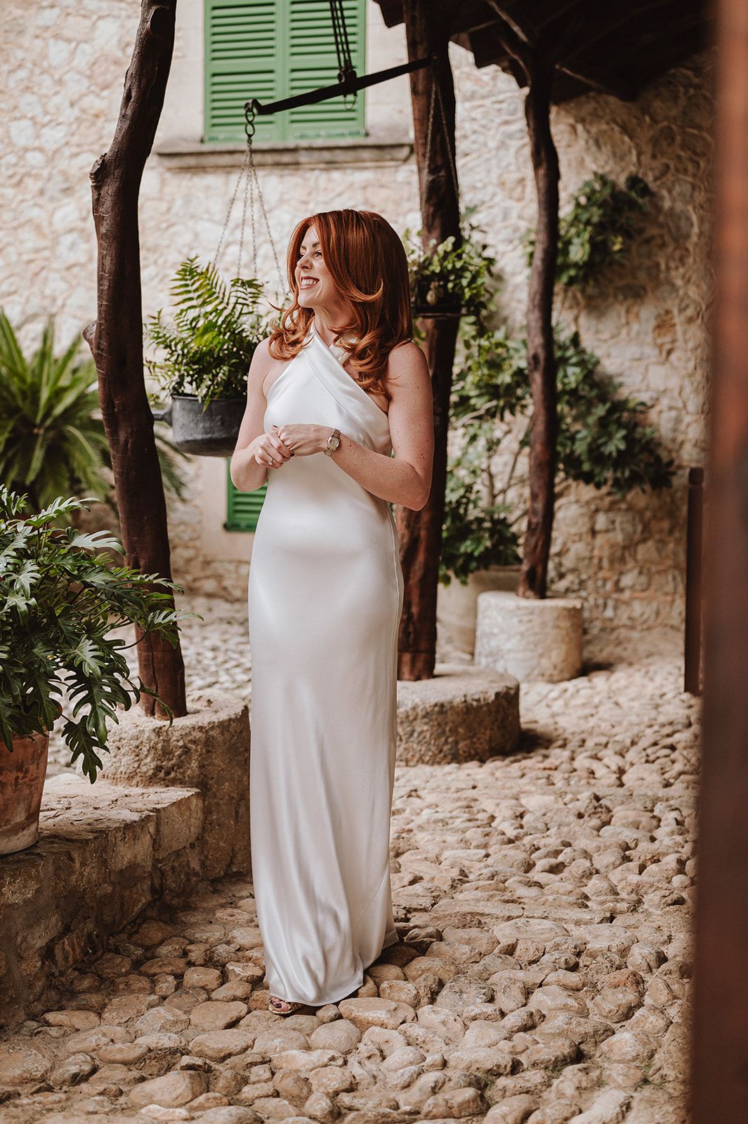 Radiant red-haired bride in a sleek white satin gown laughing in a sunlit stone courtyard.