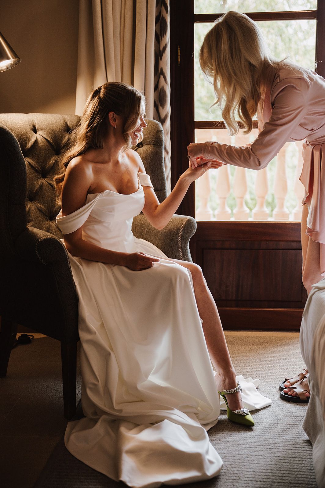 Bride in off-shoulder white gown sits in armchair as bridesmaid in pink helps put on green jeweled heels.