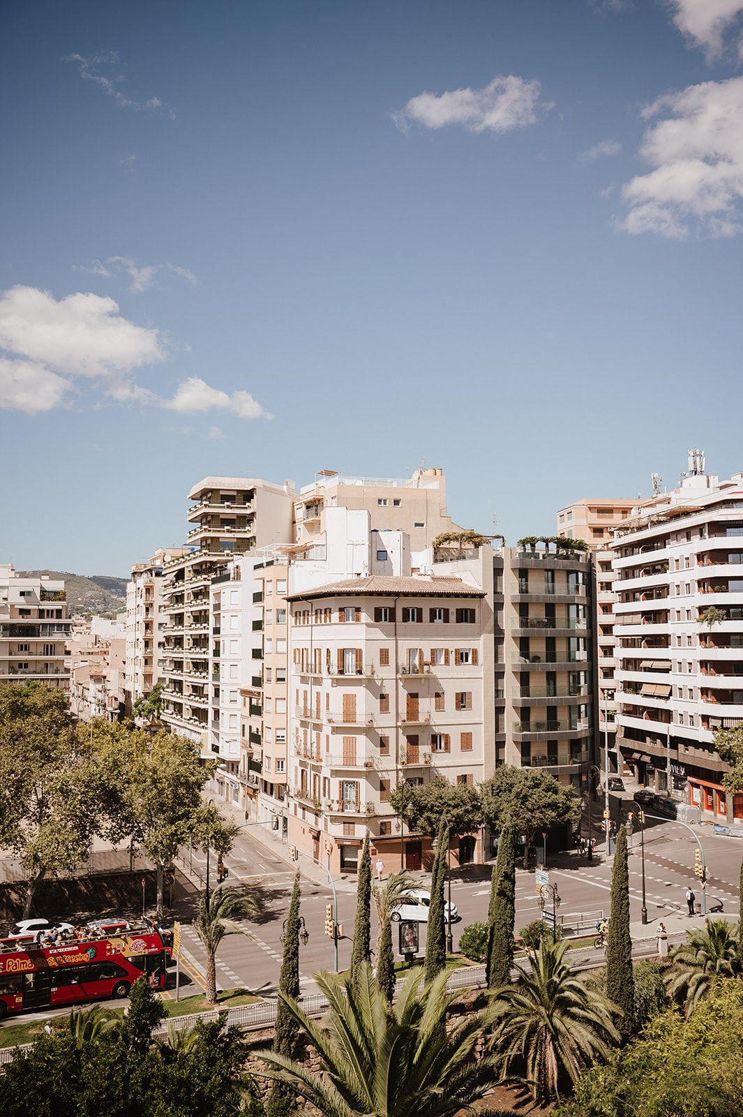 Aerial view of a sunny Mediterranean cityscape with palm trees, multi-story buildings, and a red double-decker bus.