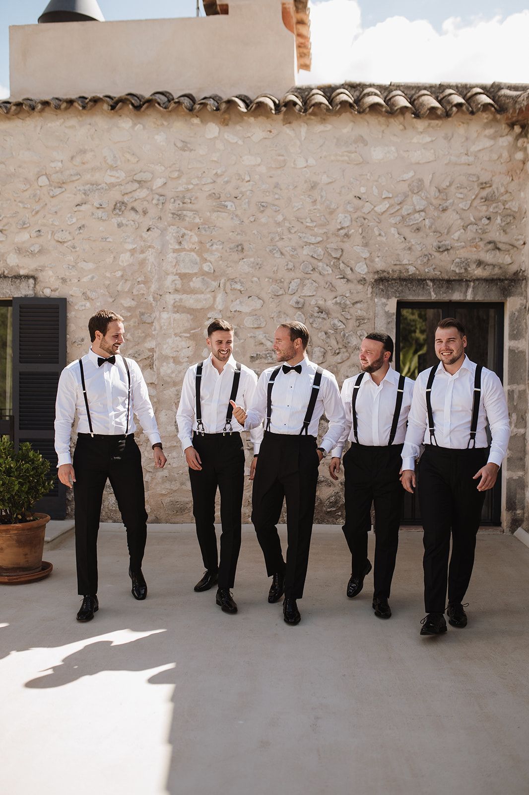 Five groomsmen in white shirts with suspenders and bow ties walking together outdoors.