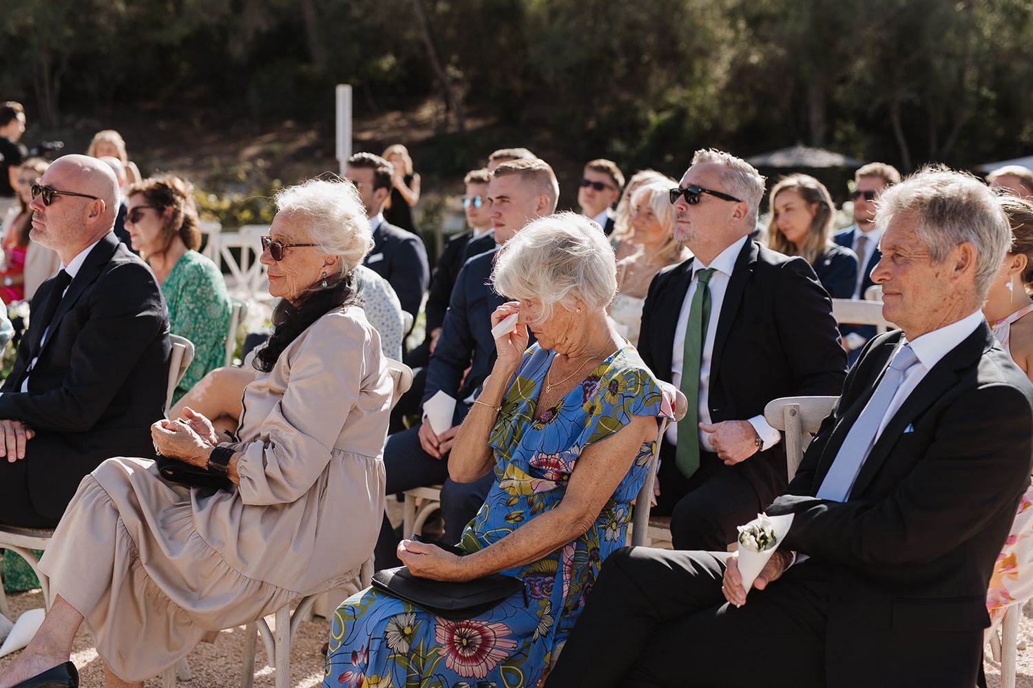 Wedding guests seated outdoors during a ceremony, with one woman wiping tears from her eyes.