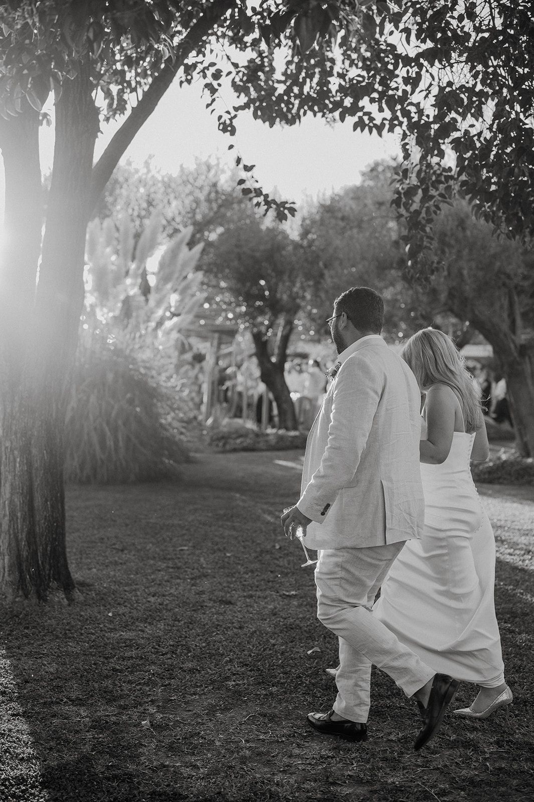 Black and white photo of bride and groom walking hand in hand through sunlit garden trees after ceremony.