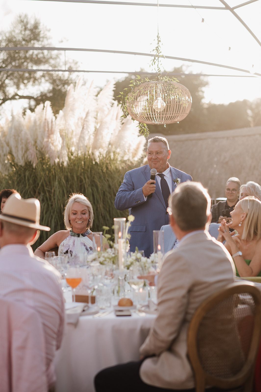 Man in blue suit giving wedding speech at outdoor reception while guests smile and listen under wicker chandelier.